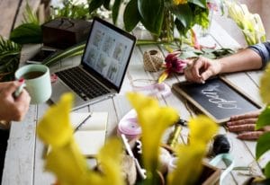 Person working on laptop at a counter in a flower shop with another person writing open on a sign