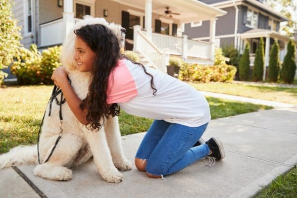 Young girl hugging a dog while kneeling on a sidewalk outside of a suburban home